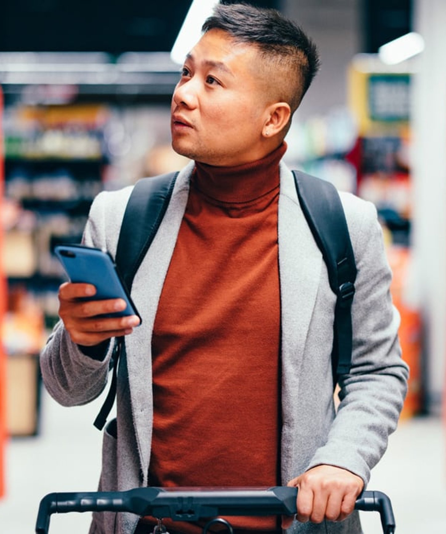 Man shopping in a grocery store, holding a smartphone and pushing a cart while looking around thoughtfully