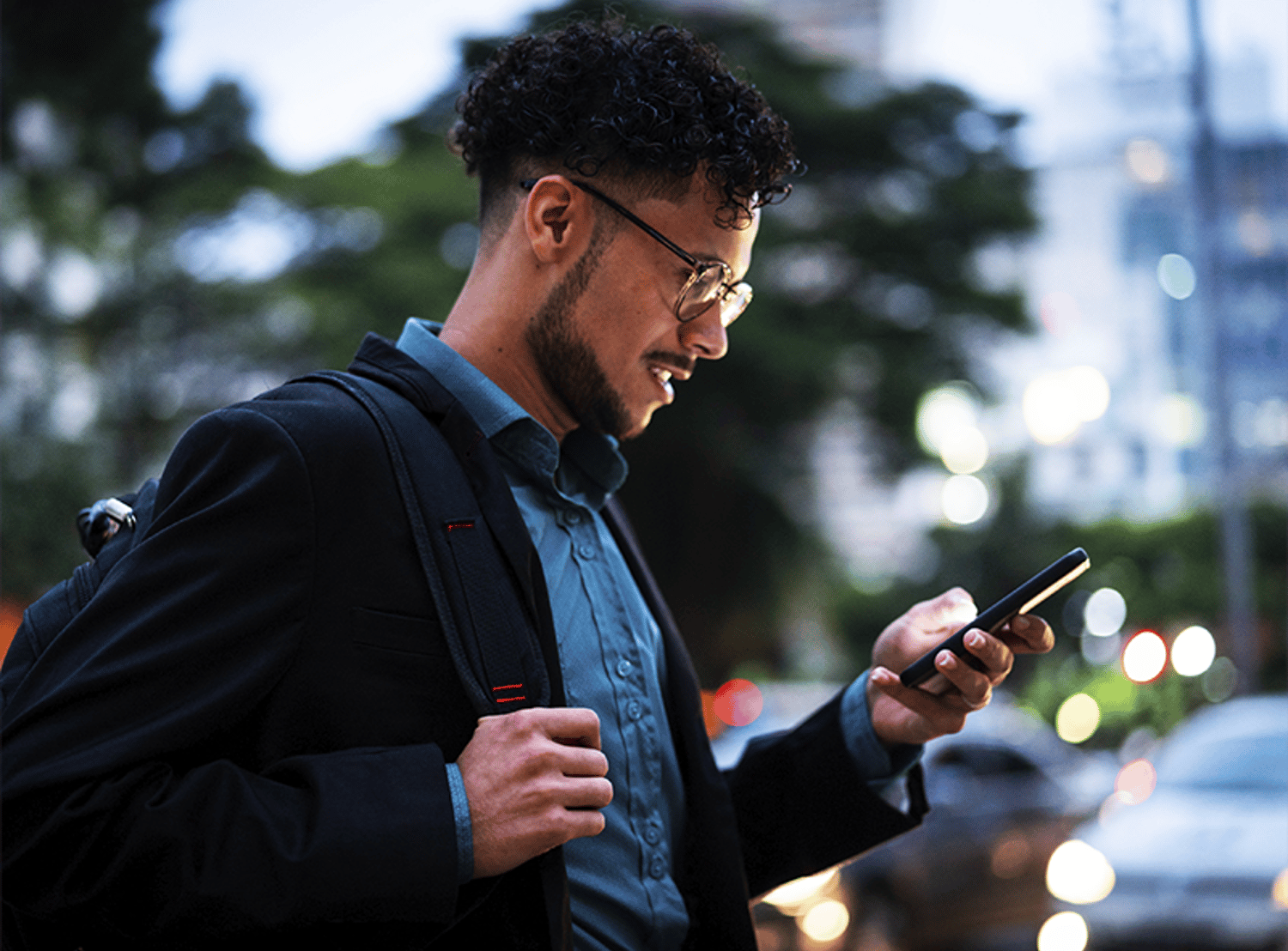 A young man with glasses and a backpack smiling while looking at his phone outdoors.