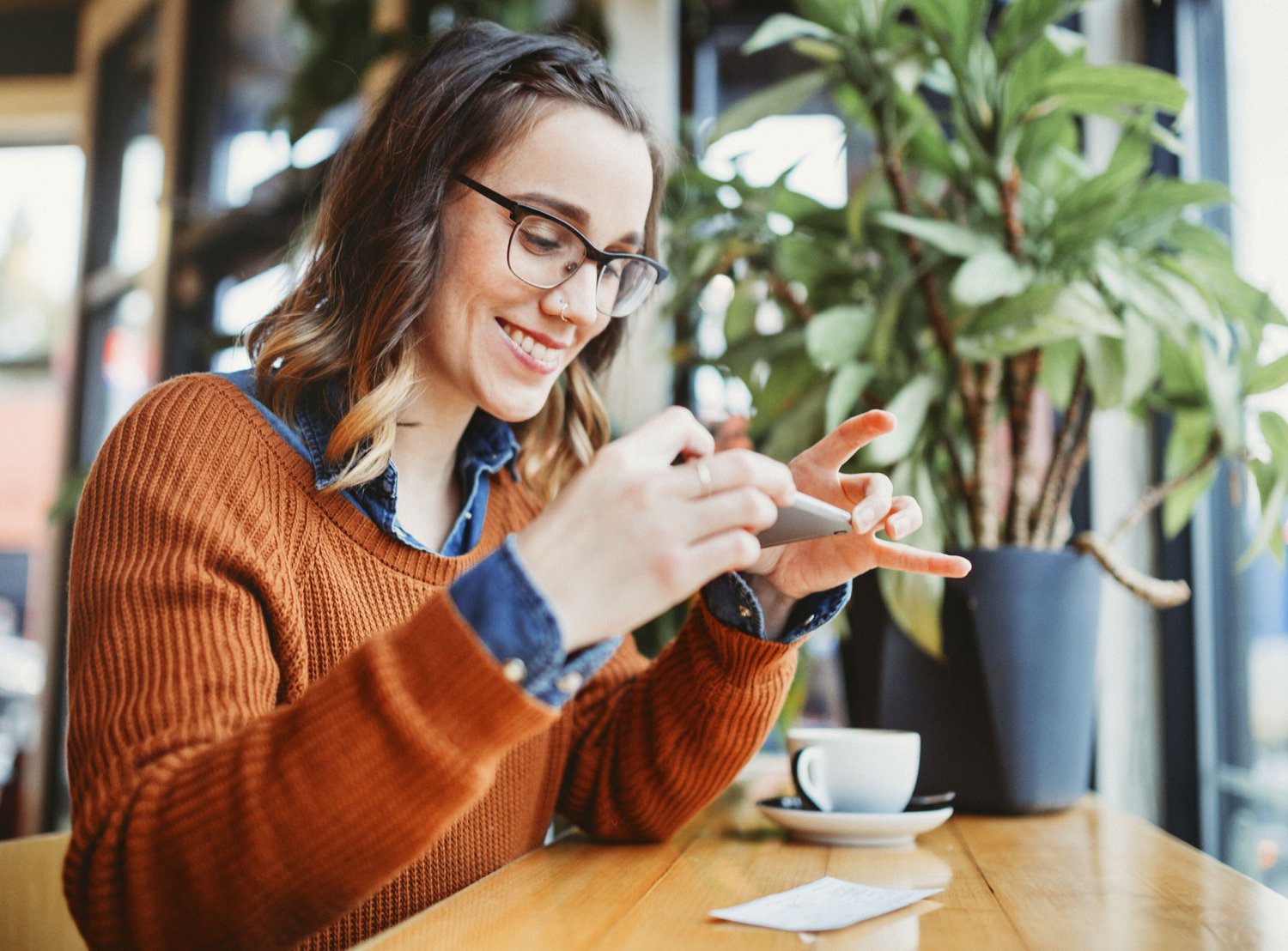 A smiling woman wearing glasses and a rust-colored sweater takes a photo of a receipt with her smartphone in a cozy café.