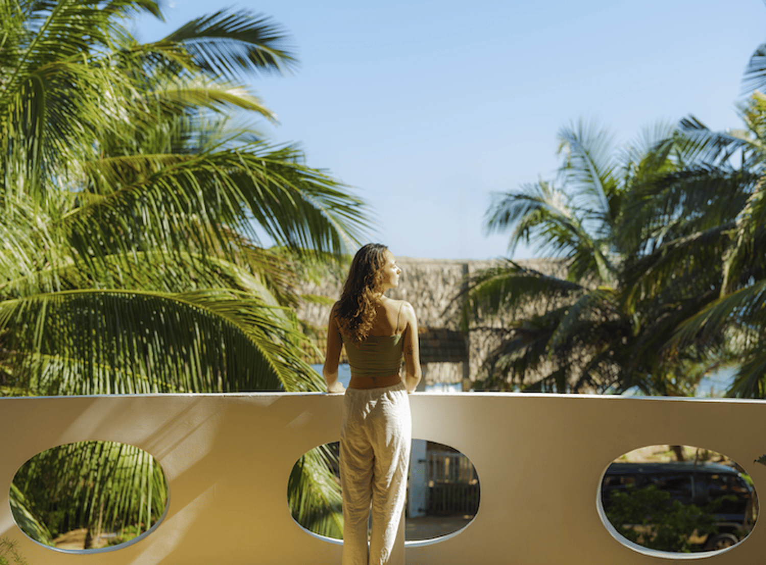 Image shows a woman standing on a balcony overlooking palm trees and basking in the sun