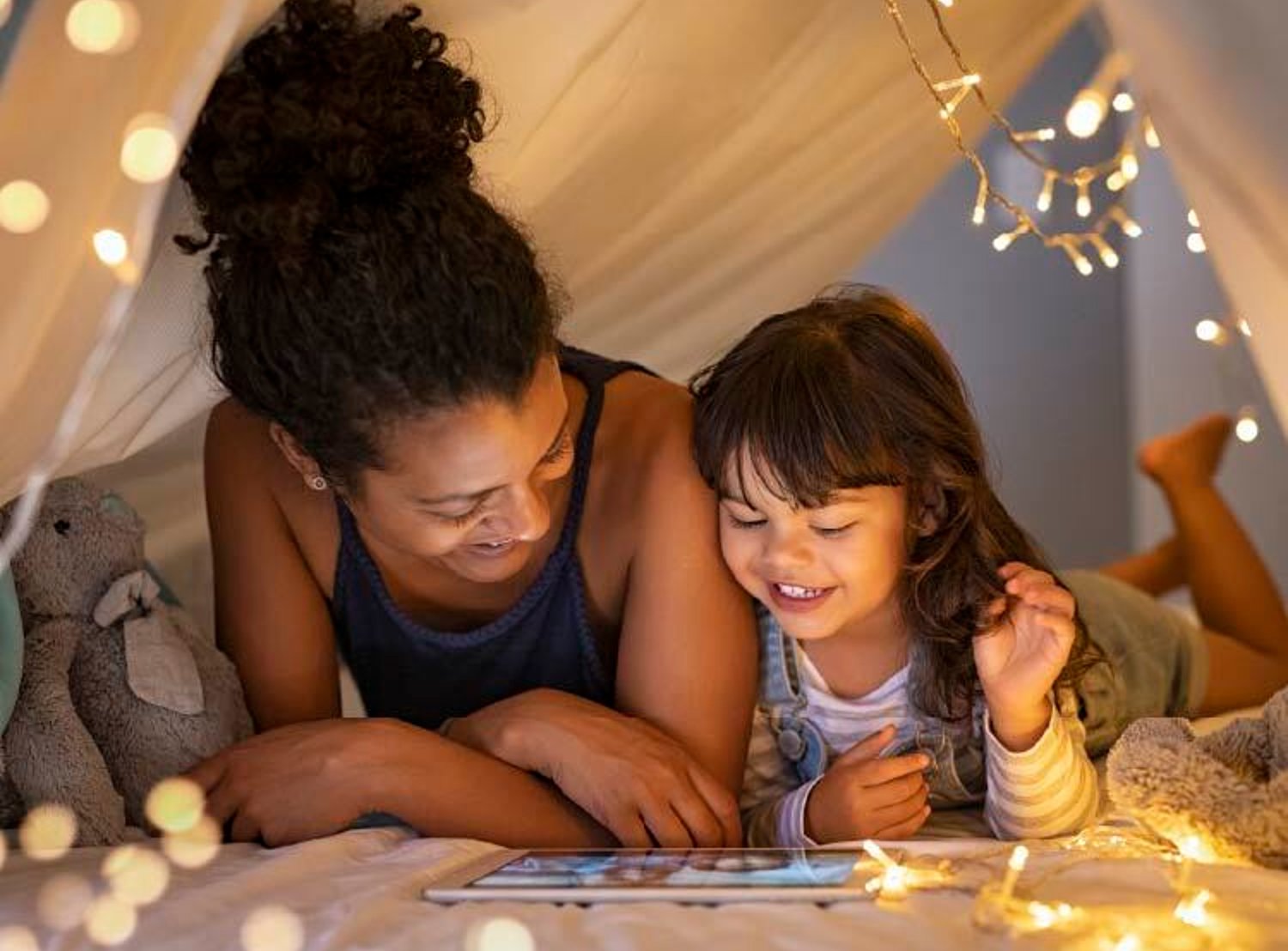 A mother and her young daughter smile while looking at a tablet together inside a cozy, decorated blanket fort with string lights.