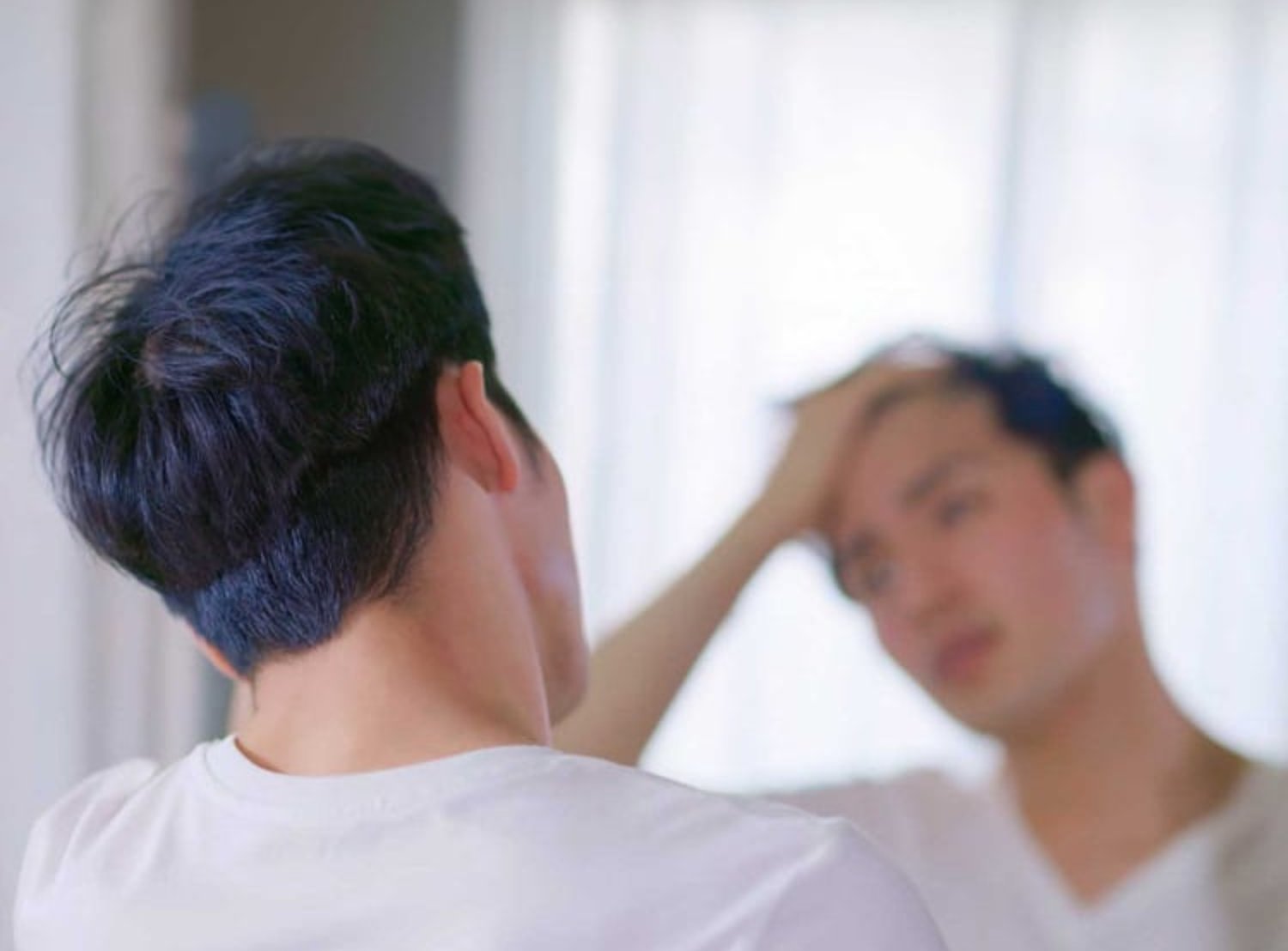 A man examines his reflection in the mirror while running a hand through his hair in a softly lit room.