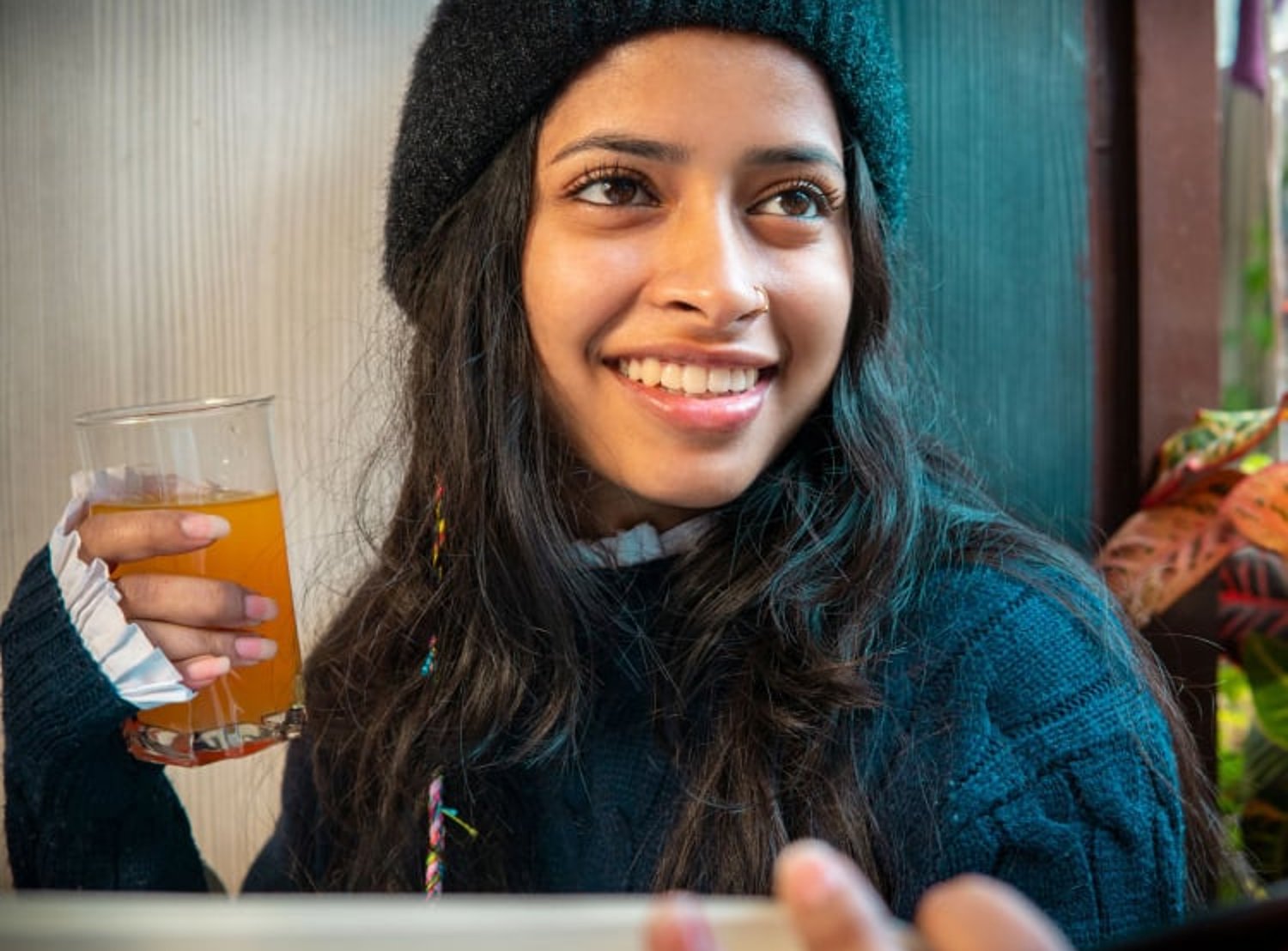 A woman smiles while holding a glass of orange-colored beverage, wearing a knit hat and sweater in a cozy indoor setting.