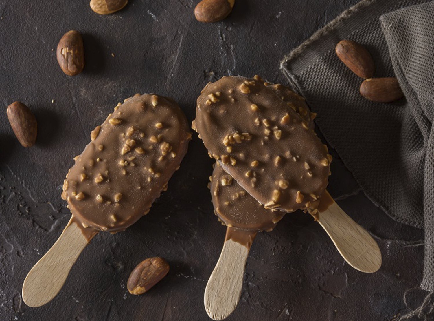 Two chocolate-coated ice cream bars with nuts on wooden sticks, placed on a dark textured surface with scattered cocoa beans.