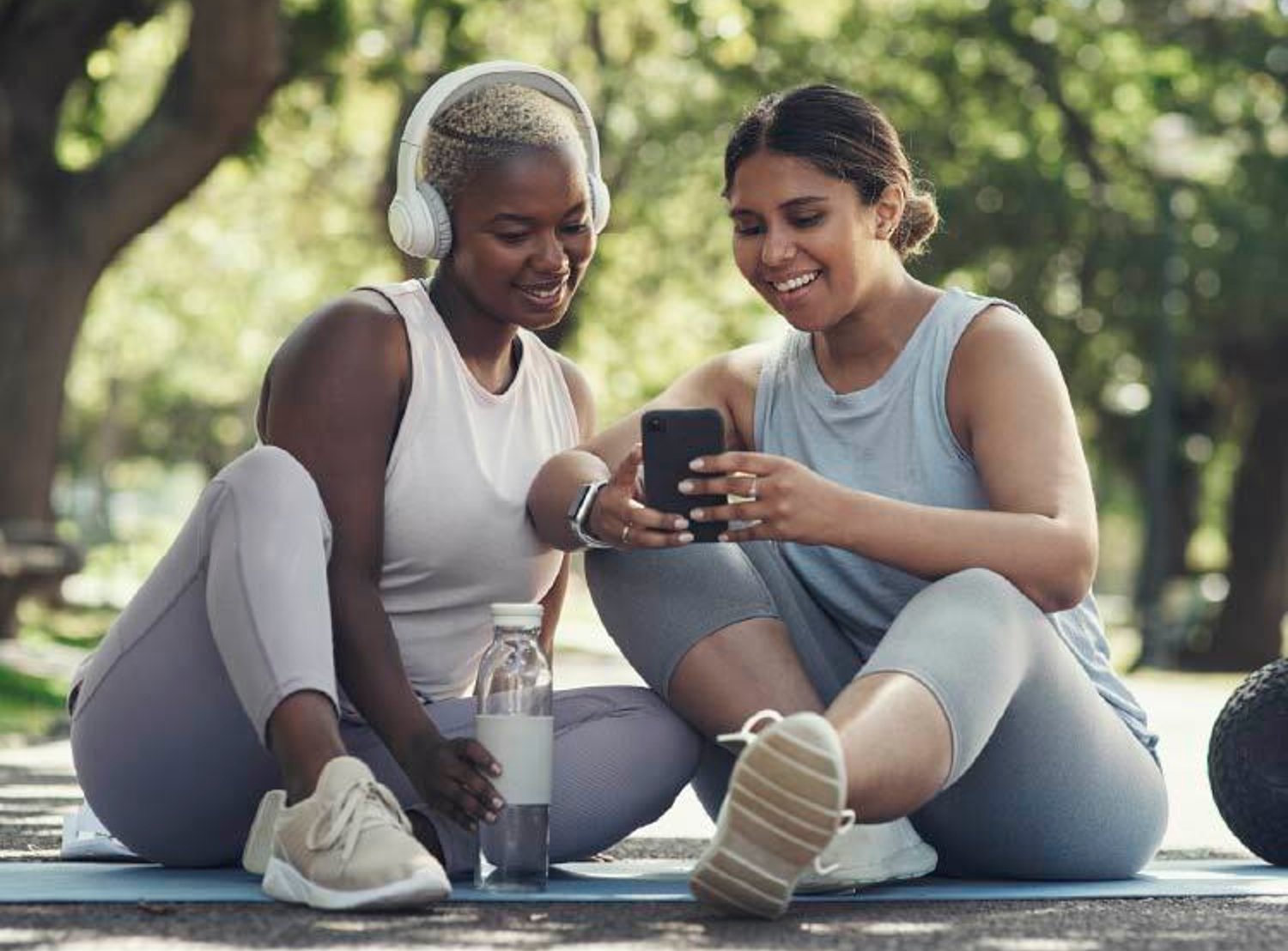 Two women in athletic wear sit outdoors, smiling while looking at a smartphone, with one wearing headphones and the other holding the phone.