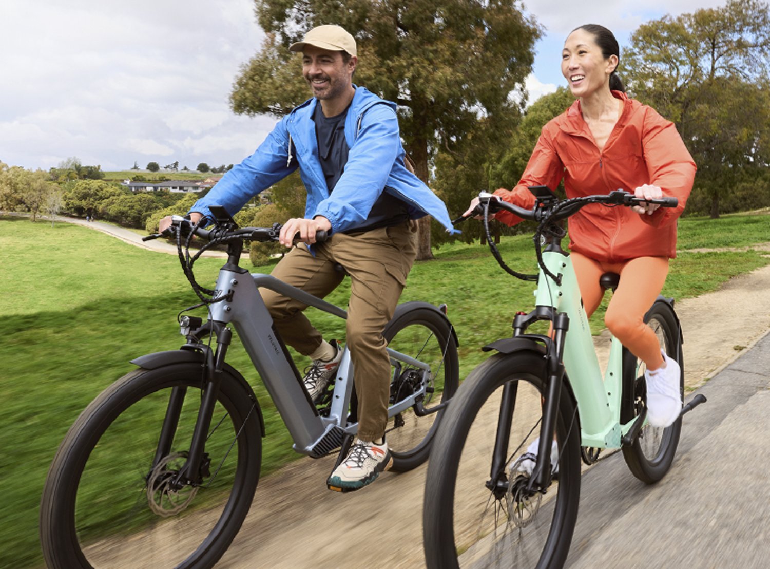 A smiling man and woman ride electric bicycles along a scenic outdoor path, dressed in sporty jackets.