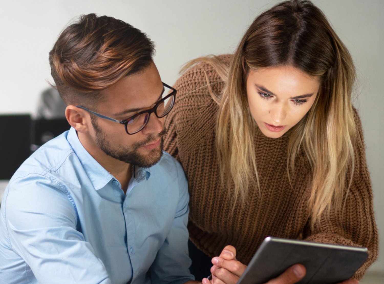 A man and woman closely collaborate while looking at a tablet, focused on a shared task in a professional setting.