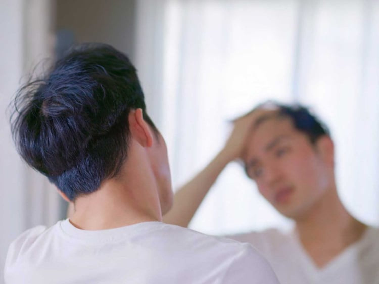 A man examines his reflection in the mirror while running a hand through his hair in a softly lit room.
