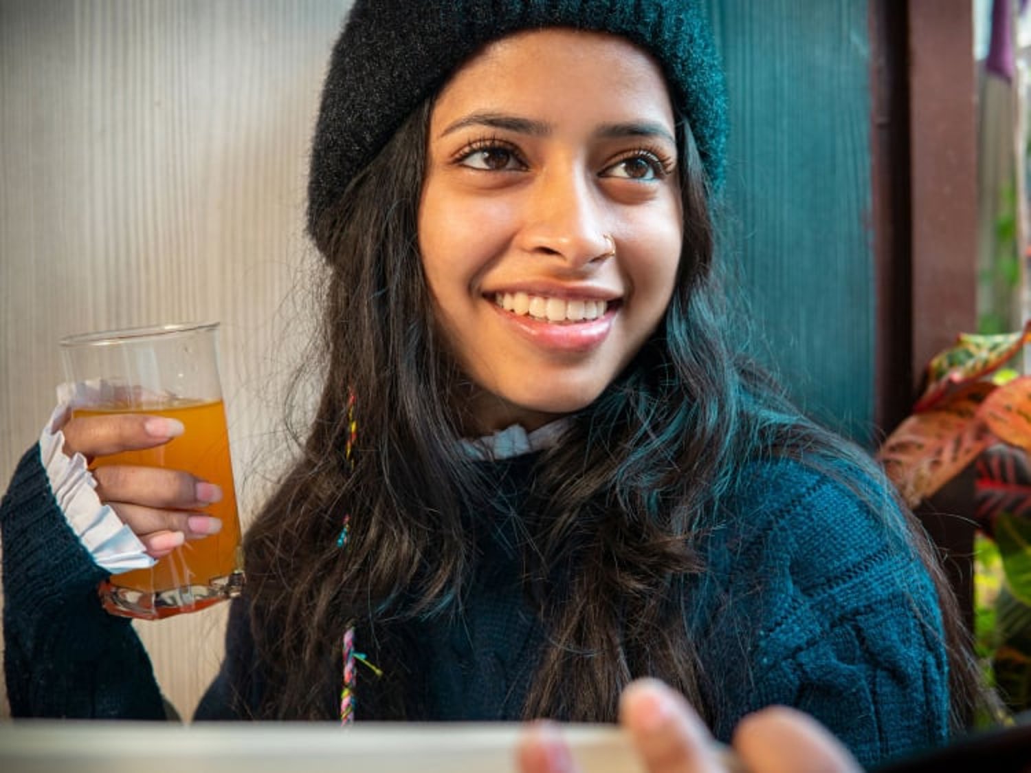 A woman smiles while holding a glass of orange-colored beverage, wearing a knit hat and sweater in a cozy indoor setting.