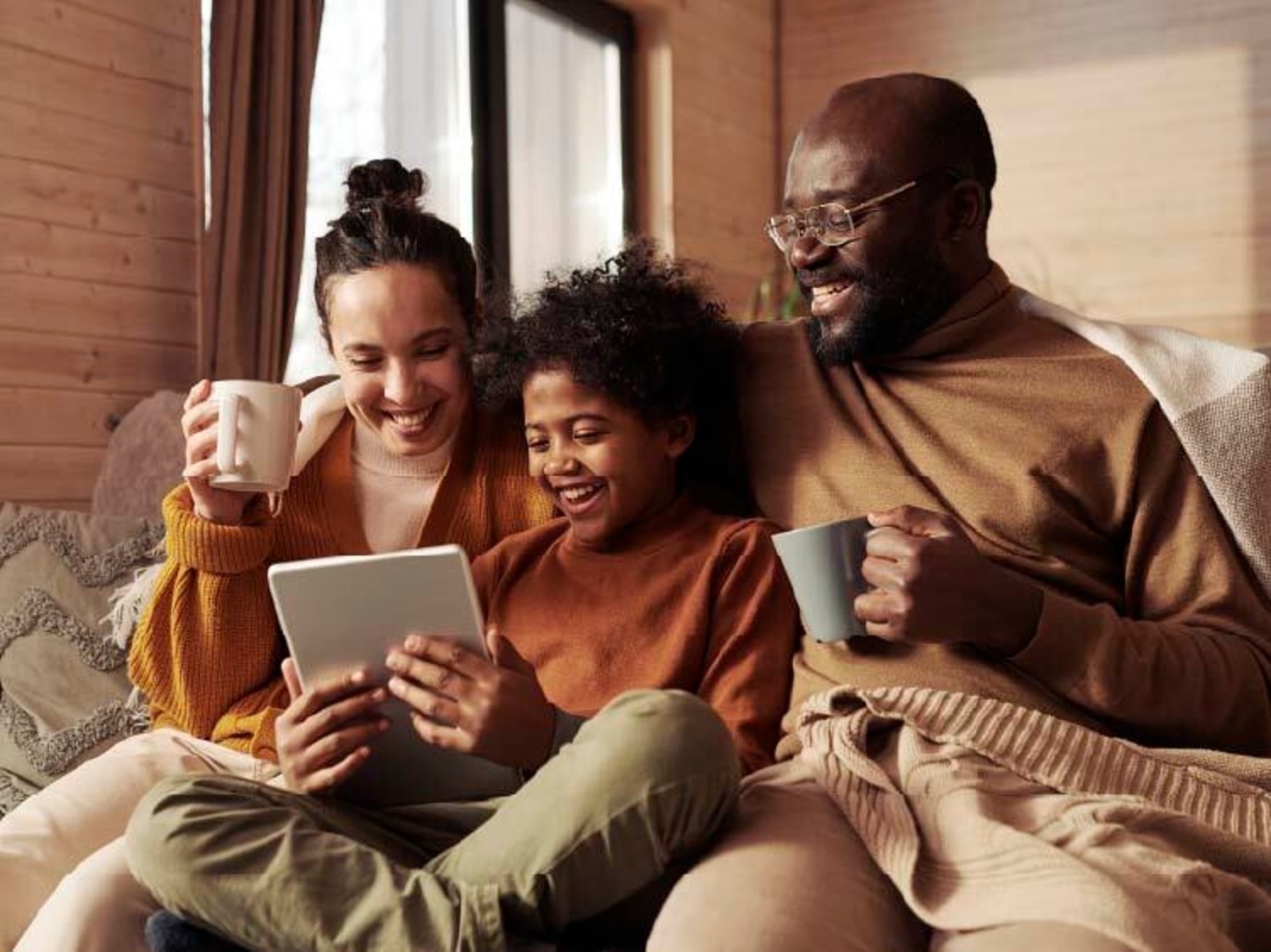 A mom, dad and child all holding each other and smiling while drinking coffees and looking at a tablet.
