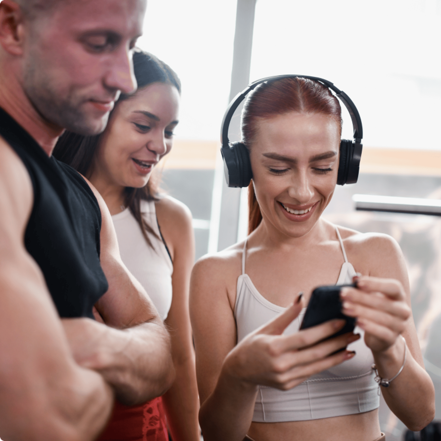 A group of three fit individuals at the gym, with a smiling woman wearing headphones showing something on her phone to the others.