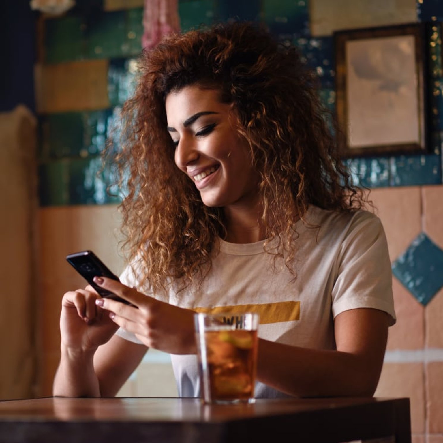 A smiling woman with curly hair sits at a café table, looking at her phone with a glass of iced tea in front of her