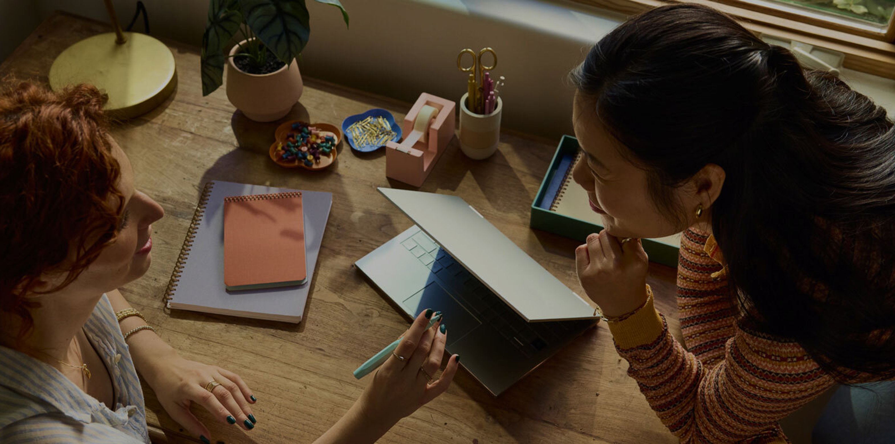 Two women sit at a wooden desk, engaged in a discussion with a partially open laptop, notebooks, and office supplies scattered around.