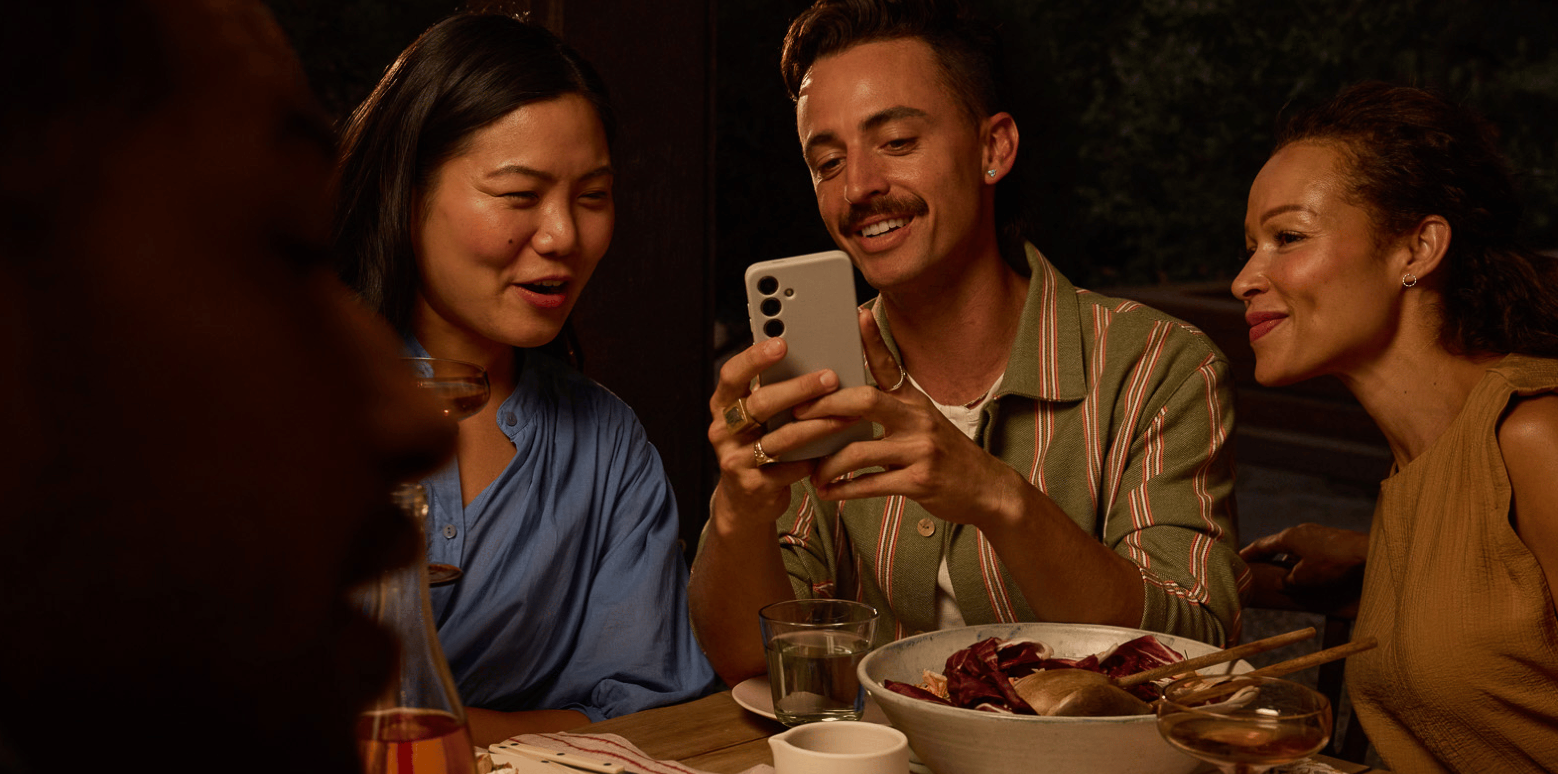 A group of friends gathers around a dinner table, sharing a moment as one person shows something on their phone, eliciting smiles and reactions.