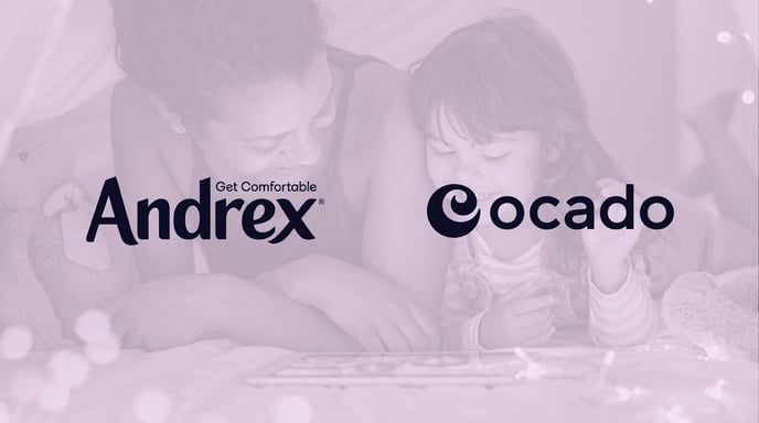 A mother and her young daughter smile while looking at a tablet together inside a cozy, decorated blanket fort, overlaid with the Andrex and Ocado logos and the tagline "Get Comfortable."