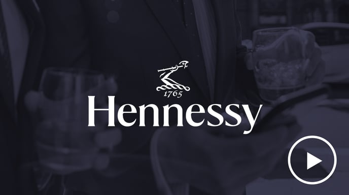 Two men in suits holding glasses of Hennessy while looking at a smartphone, overlaid with the Hennessy logo and a play button.