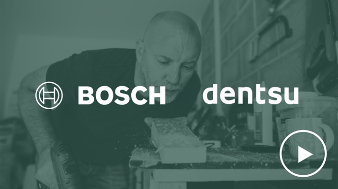 A bald man with a beard, wearing a navy blue t-shirt, is using a power drill on a wooden plank while focusing intently in a workshop setting with Bosch and Dentsu logos placed over