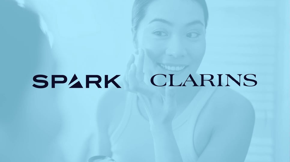 Woman looking in the mirror and smiling, while doing her skincare, with a blue overlay over and Spark + Clarins logos over the top.