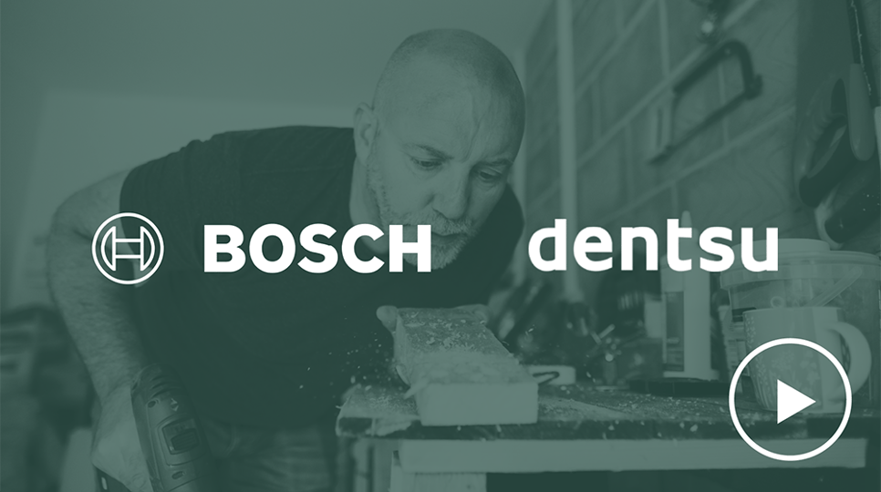 A bald man with a beard, wearing a navy blue t-shirt, is using a power drill on a wooden plank while focusing intently in a workshop setting with Bosch and Dentsu logos placed over